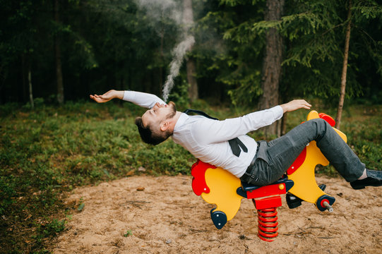 Adult Serious Man In Business Clothes Riding Children Metal Horse Attraction With Spring On Playground. Odd Person Blows Out Clouds Of Thick Smoke. Strange Posing With Arms Apart. Weird Behavior.