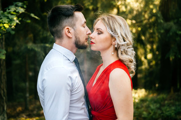 Closeup of couple of lovers kissing with smoke outdoor.  Businessman and young blonde girl with red lips, curly hair and red dress with decolette. Smokers in forest in summer sunny day. Sensual moment