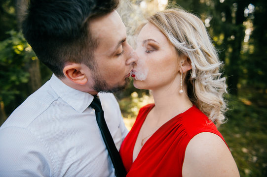 Closeup of couple of lovers kissing with smoke outdoor.  Businessman and young blonde girl with red lips, curly hair and red dress with decolette. Smokers in forest in summer sunny day. Sensual moment