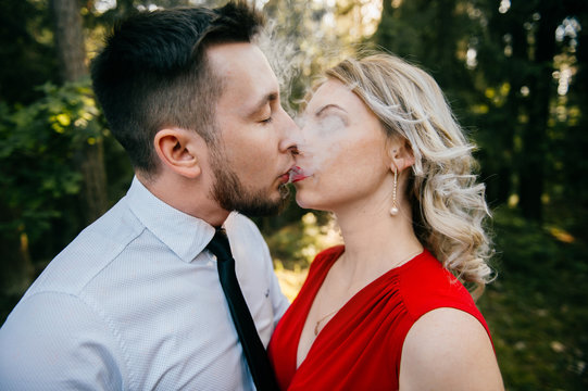 Closeup of couple of lovers kissing with smoke outdoor.  Businessman and young blonde girl with red lips, curly hair and red dress with decolette. Smokers in forest in summer sunny day. Sensual moment