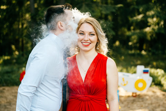 Smoking man in business clothes kissing young blonde girl with red lips, dress with decolette.  Clouds of thick smoke. Couple of lovers outdoor.  Smiling and laughing woman. E-cigarette.