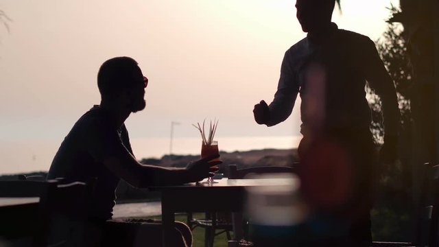 Silhouette of a young man in the bar at sunset. The waiter brings him a drink