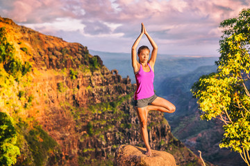 Yoga girl doing tree pose on mountain nature landscape in Waimea Canyon, Kaui, Hawaii doing Vriksasana one leg standing. Woman practicing meditation in sunset, health and wellness concept.