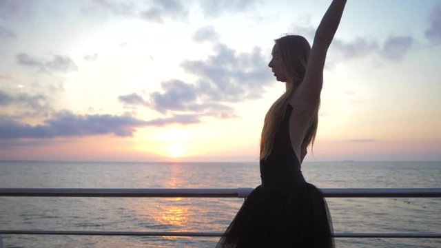 Beautiful scene of a dancing ballerina in black ballet tutu and pointe on embankment above ocean or sea beach at sunrise or sunset. Young beautiful blonde woman with long hair practicing stretching