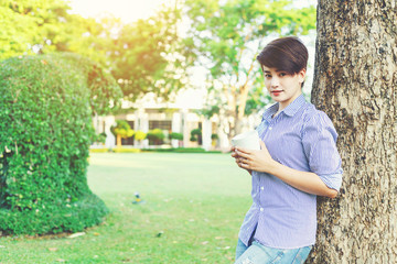 Beautiful short hair woman standing under giant tree and holding a favorite beverage in white cup between her outdoor working business in the park.