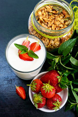 A set of a product for a delicious healthy breakfast: granola, yogurt, fresh strawberries, green mint leaves on a black background. Proper nutrition. Dietary menu. Vertical view.