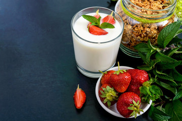 A set of a product for a delicious healthy breakfast: granola, yogurt, fresh strawberries, green mint leaves on a black background. Proper nutrition. Dietary menu.