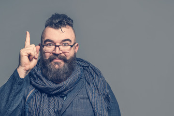 Adult bearded man with glasses and scarf showing forefinger up on a gray background. Isolated
