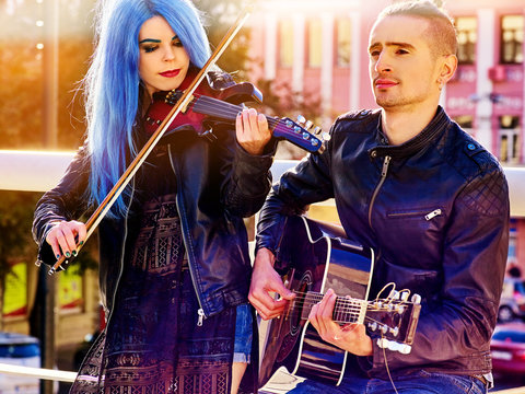 Playing viola woman and man perform music on violin and guitar in city outdoor. Girl with blue hairstyle and eyebrows performing jazz on urban street. Couple of musicians in love makes living.