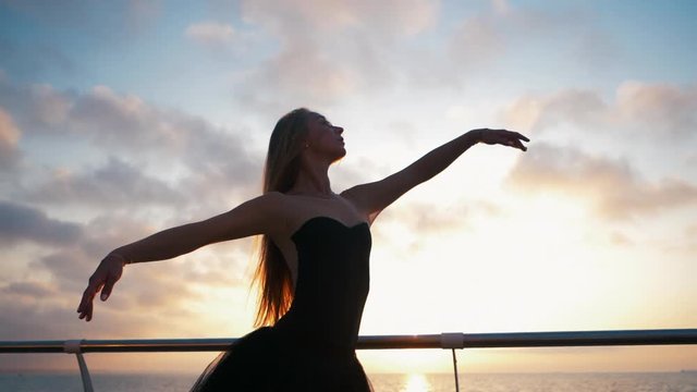 SLOW MOTION. Beautiful ballerina performing pirouette. Blonde woman in black ballet tutu on embankment above ocean or sea beach at sunrise. Female dancing with arms.