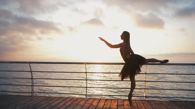 Beautiful scene of a dancing ballerina in black ballet tutu and pointe on embankment above ocean or sea beach at sunrise or sunset. Silhouette of young beautiful blonde woman with long hair practicing