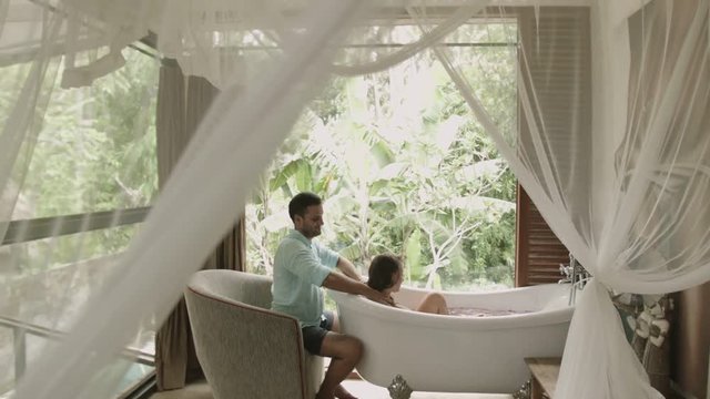 Woman takes the bathroom in the room with panorama windows overlooking the tropical garden. Man strokes woman's skin and doing massage