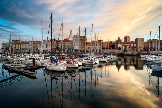 Sunrise landscape scenery of Gijon, Spain, Europe. Beautiful reflection on calm sea water of boats, buildings, sky at dusk at touristic cultural travel destination.