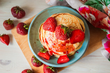 Beautiful appetizing pancakes and strawberries on a wooden table on a white brick wall background. Pancakes with jam, jam and fruits. Flowers in the vase and homemade food.   