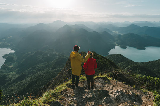 Woman in red jacket and male with yellow coat looking over a valley with mountains, forest and lake in Germany in the morning and having fun