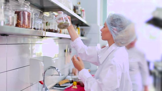 Cook is looking for on the shelf a jar with spices. The woman take a jar with spices and smiles.