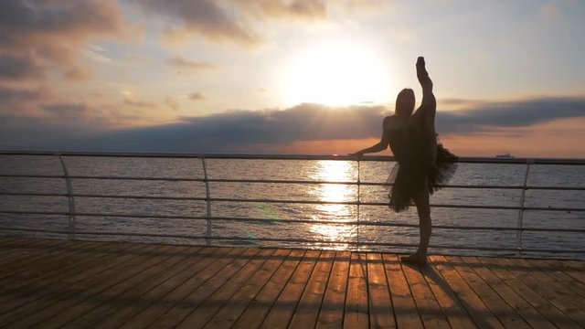 Silhouette of a dancing ballerina in black ballet tutu and pointe on embankment above ocean or sea beach at sunrise or sunset. Silhouette of young beautiful blonde woman raises a leg up above head