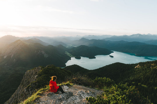 Woman in red jacket looking over a valley with mountains, forest and lake in Germany in the morning