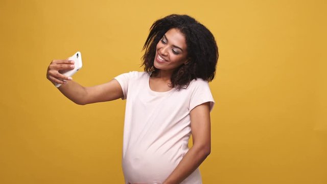 Happy afro american pregnant woman waving her hand while having video call conversation isolated over yellow background