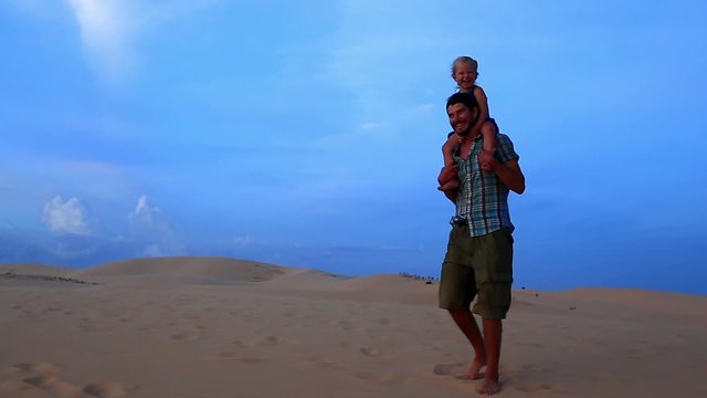 Father Jumps with Small Girl on Shoulders against Dunes Blue Sky