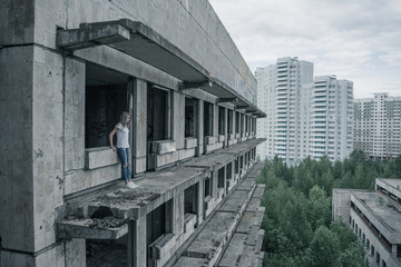 A young girl in a white T-shirt and blue jeans is standing on the edge of a ruined balcony in a ruined building early in the morning.