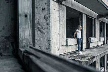 A young girl in a white T-shirt and blue jeans is standing on the edge of a ruined balcony in a ruined building early in the morning.