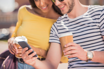 Cheerful young multiethnic couple with coffee cups using smartphone in city