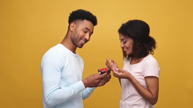 Smiling young african man giving a present box to his girlfriend isolated over yellow background