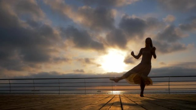 Jumping ballerina in beige silk dress and pointe on embankment above ocean or sea beach at sunrise or sunset. Silhouette of young beautiful blonde woman with long hair practicing stretching and