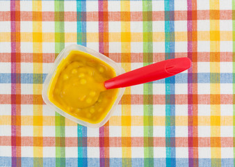 Macaroni and cheese baby food in a container with a red plastic spoon in the food atop a colorful place mat.