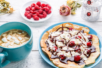 Homemade pancakes or Russian pancakes with chocolate sauce, whipped cream and raspberries on a plate on a white wooden background.
