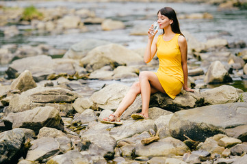 Young lonely tourist cutie girl sitting on stone on shore of river in mountains in wild terrain and satisfying thirst drinking water from glass. Traveler resting at nature. Fluid balance. Heat weather