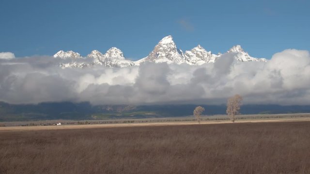 AERIAL: Majestic Grand Teton mountain peaks raising above the foggy clouds in dry Jackson Hole valley in Wyoming, United States. Pointy Rocky Mountains peaking above dense fog on sunny meadow field
