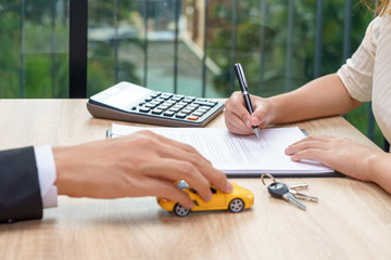 Woman signing car loan agreement contract with car key and calculator on wooden desk.