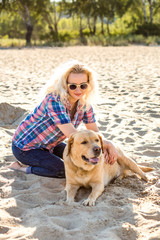 Portrait of young beautiful woman in sunglasses sitting on sand beach with golden retriever dog. Girl with dog by sea.