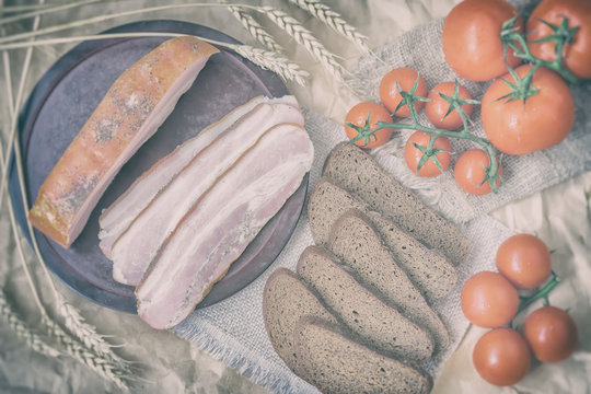 Rustic style of fresh organic food. Quick tasty snack. Slices of bacon, rye black bread, ripe red tomatoes on napkin from burlar. Top view. Vintage backdrop