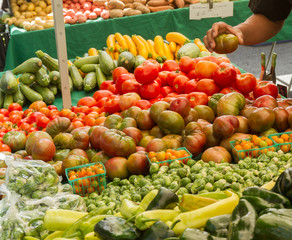 Farmer's market produce for sale at stand, with person holding an heirloom tomato. Cherry tomatoes, brussel sprouts, zucchini, squashes, gypsy peppers and bell peppers are visible.