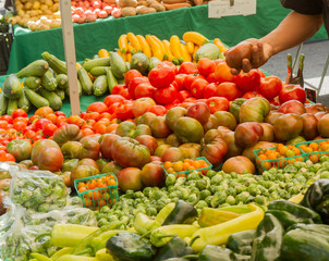 Farmer's market produce for sale at stand, with person holding an heirloom tomato. Cherry tomatoes, brussel sprouts, zucchini, squashes, gypsy peppers and bell peppers are visible.