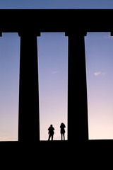 Fototapeta premium Silhouette of tourists standing amongst huge pillars of a monument taking photos of the sunset. Scottish National Monument, Edinburgh, Scotland, UK