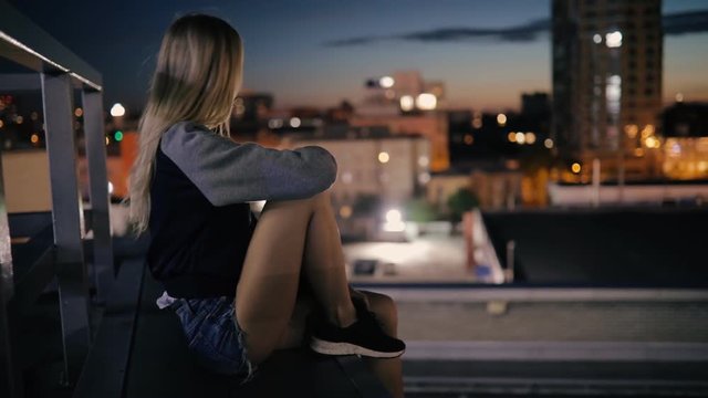 Young woman sitting on rooftop in city at night and looks to cityscape