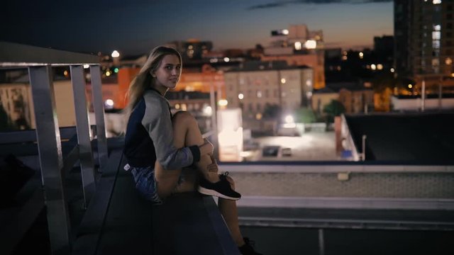 Young woman sitting on rooftop in city at night and looks to cityscape