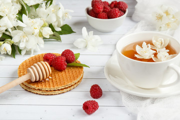 Tea Cup with Jasmine flowers and thin waffles with honey and raspberry on a light wooden background
