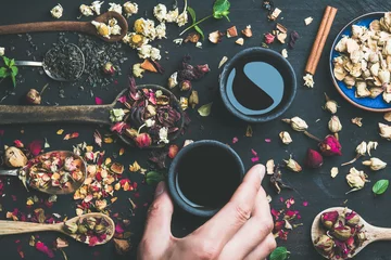 Papier Peint photo Theé Chinese black tea in black stoneware cups, man's hand holding one cup and wooden spoons with dry herbs, flower buds and leaves over black wooden background, top view, horizontal composition