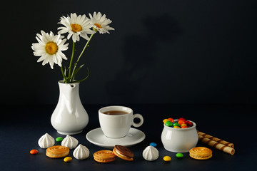 Cup with tea, teapot, sweets and white daisies on black background