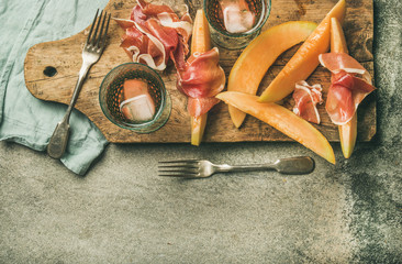 Summer wine party set. Flatlay of Wine snack Prosciutto ham and melon and glasses of rose wine with ice on rustic wooden serving board over grey concrete background. Top view, copy space
