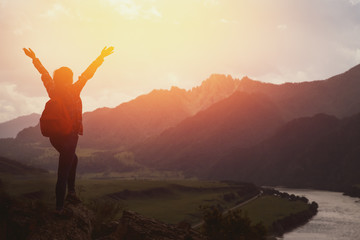 tourist girl in a plaid shirt and with a backpack standing on top of the mountain and having fun against the backdrop of mountains, rivers. The setting sun. Concept active rest. Space for text