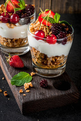 Healthy breakfast. Summer berries and fruits. Homemade Greek yoghurt with granola, blackberries, strawberries, cherries and mint. On wooden and stone black background table, in glasses. Copy space