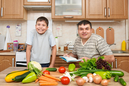 Father and child reading cooking book and choice dishes. Happy family having fun with fruits and vegetables in home kitchen interior. Healthy food concept.
