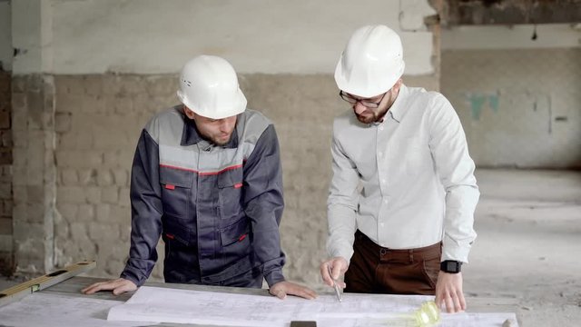 Two engineers in protective helmets working together in construction area. Inspector and foreman standing in building site and investigating structural drawing. Builders discussing current project.
