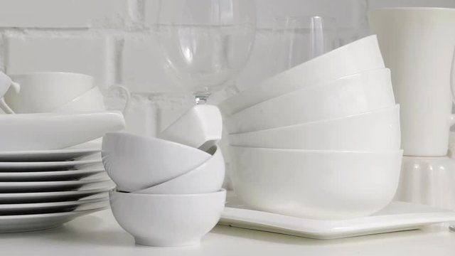 Stacked white porcelain dishes, plates, cups, saucers, mugs in restaurant kitchen interior. Slow pan. White brick wall background.	 Selective focus.